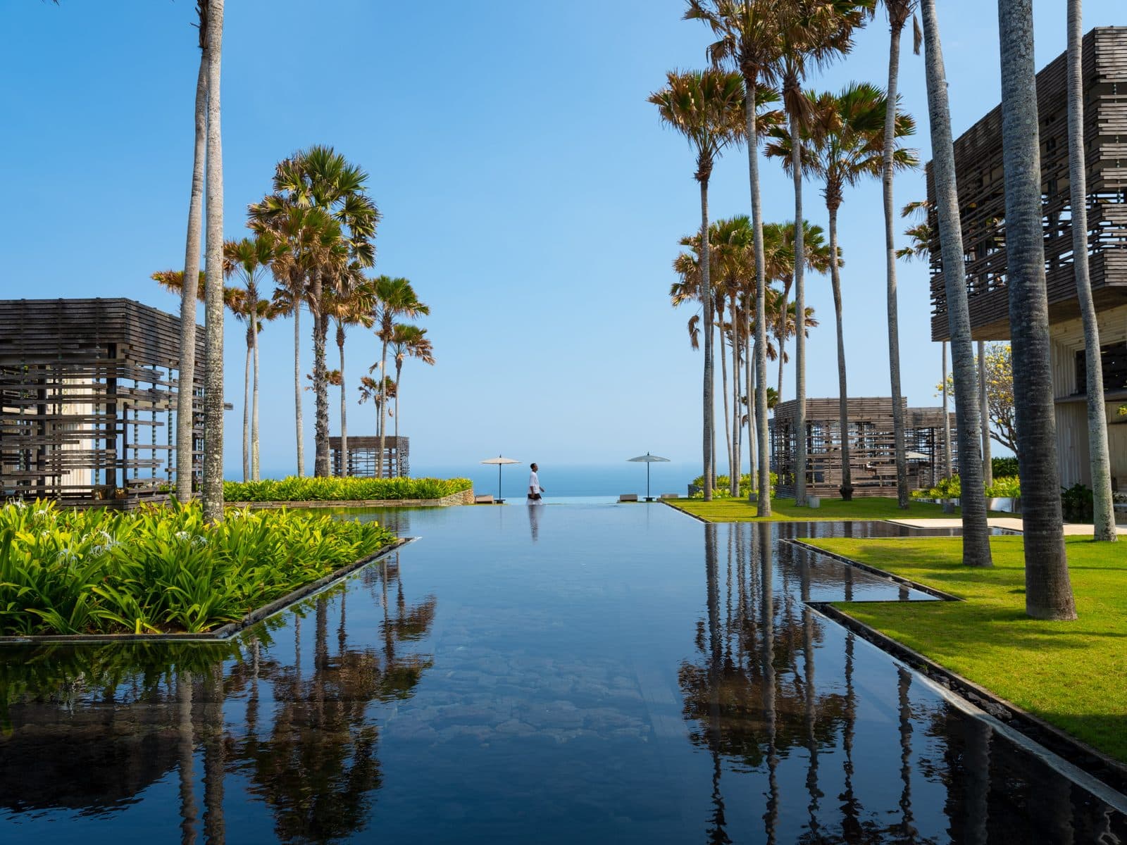 Alila Villas Uluwatu: a long reflecting pool between lawns and tall palms leads towards the ocean horizon, with wooden lattice pavilions at the sides