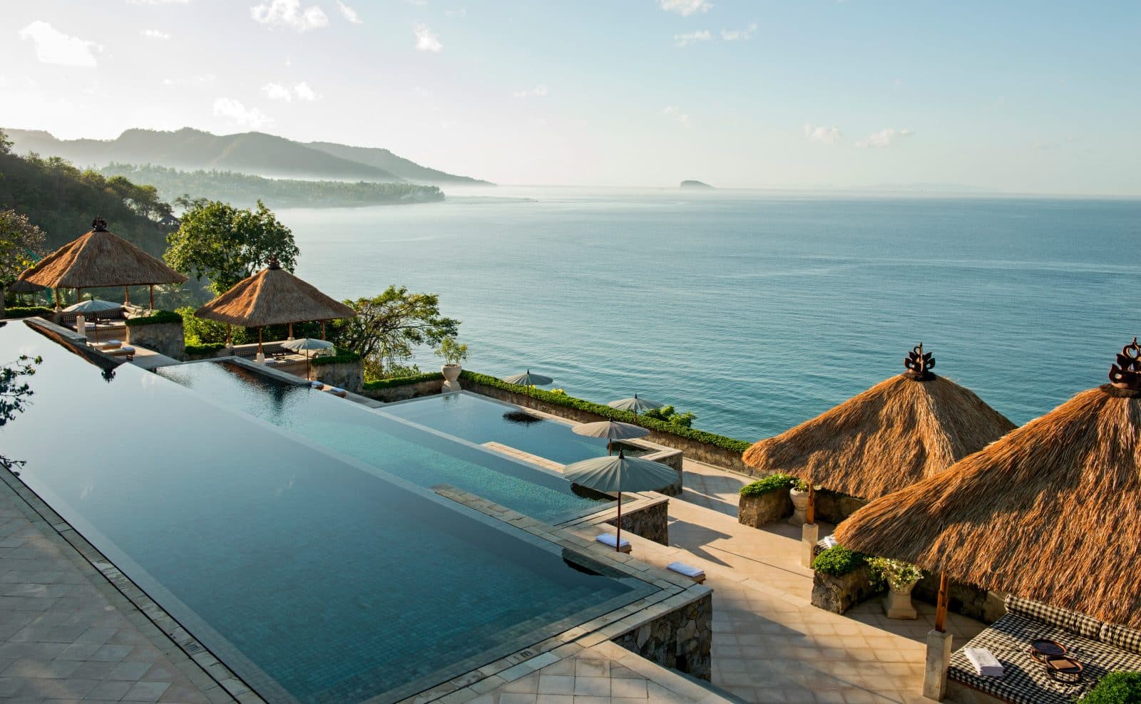 Amankila's three-tier pool above the Lombok Strait: thatched pavilions on terraces, umbrellas and an island at sea in the distance