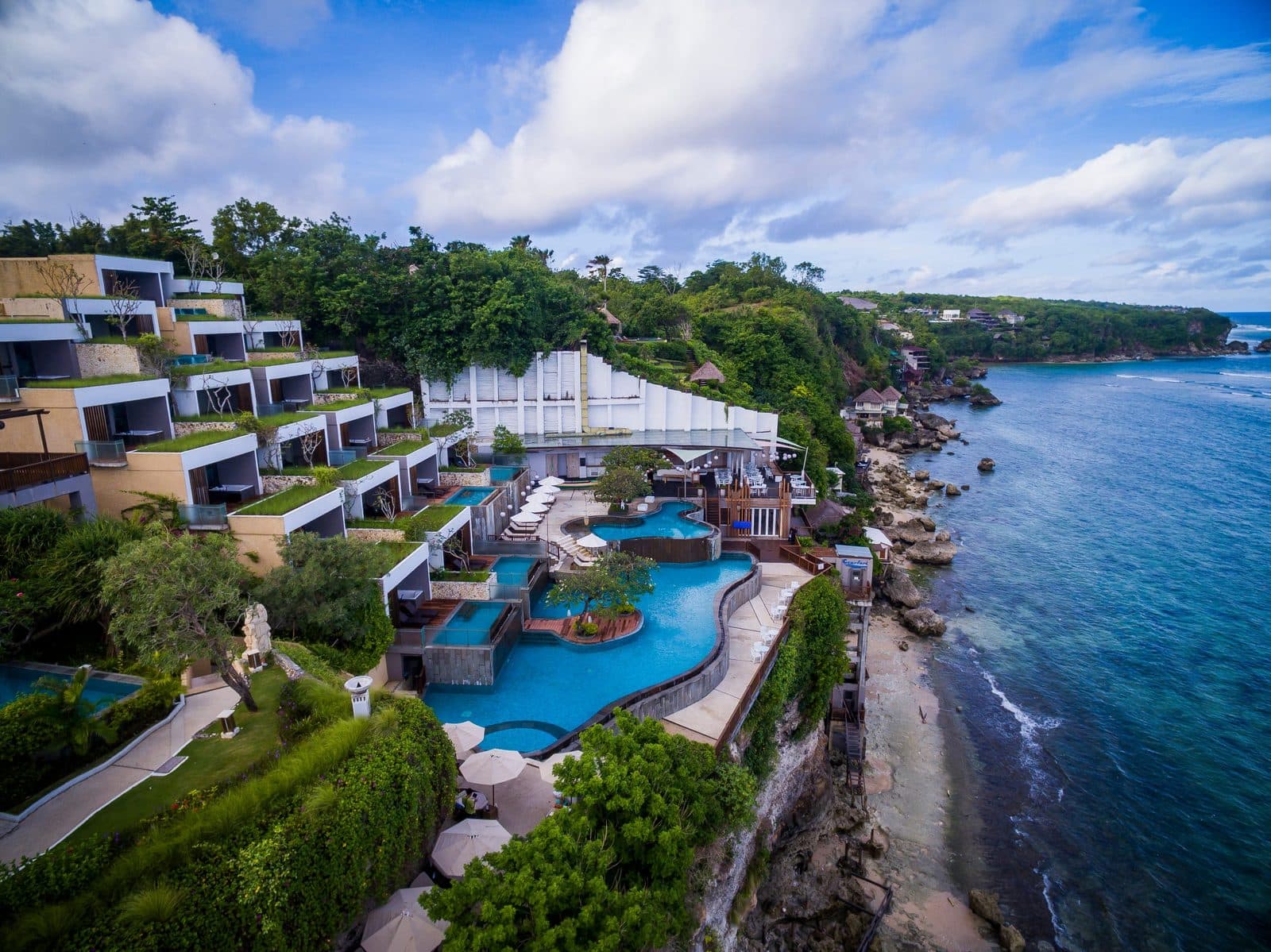 Aerial view of Svanata Uluwatu: buildings stepped down the slope, a multi-level pool above the cliff and a rocky ocean shore