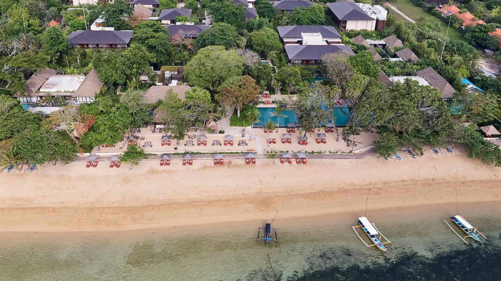 Aerial view of Andaz Bali and Sanur Beach: roofs among trees, a pool by the shore, rows of loungers and jukung boats in the shallows