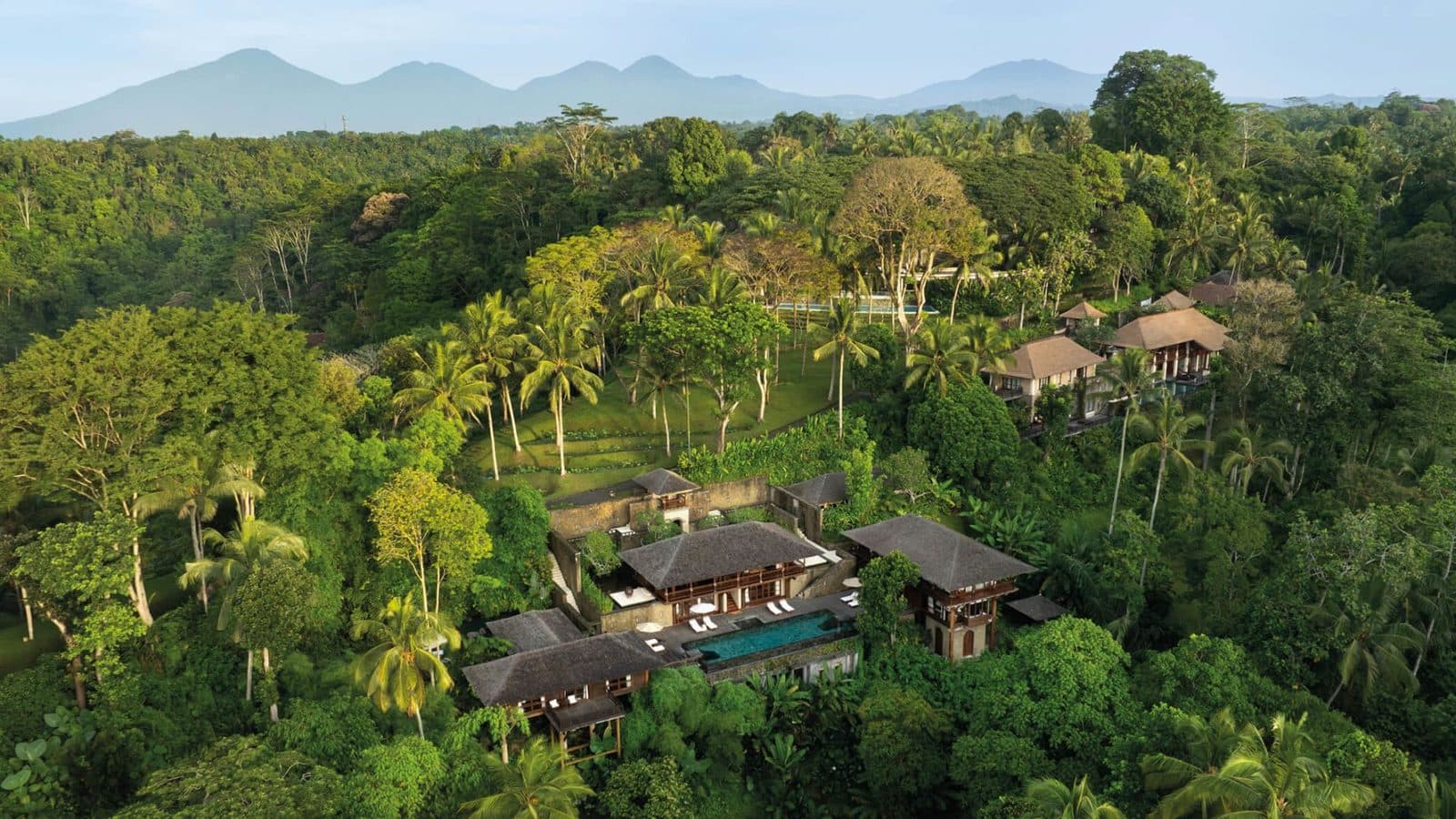 Aerial view of Tirta Ening residence: dark-roofed houses and a pool among forest, with mountains on the horizon