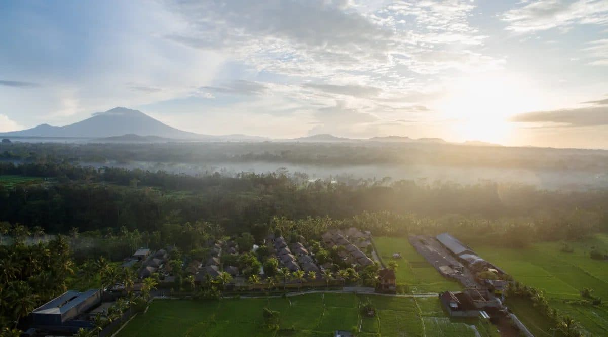 Aerial view of HOSHINOYA Bali at sunrise: villa roofs among palms and rice fields, mist over the valley and a volcano on the horizon