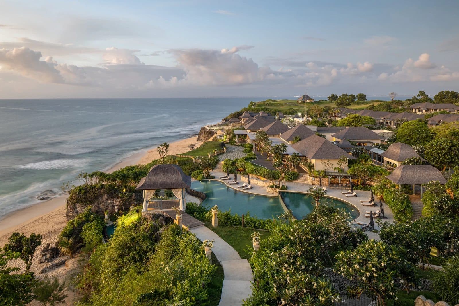 Jumeirah Bali at sunset: dark-roofed villas in a garden above Dreamland Beach, a free-form pool and a cliff-edge gazebo
