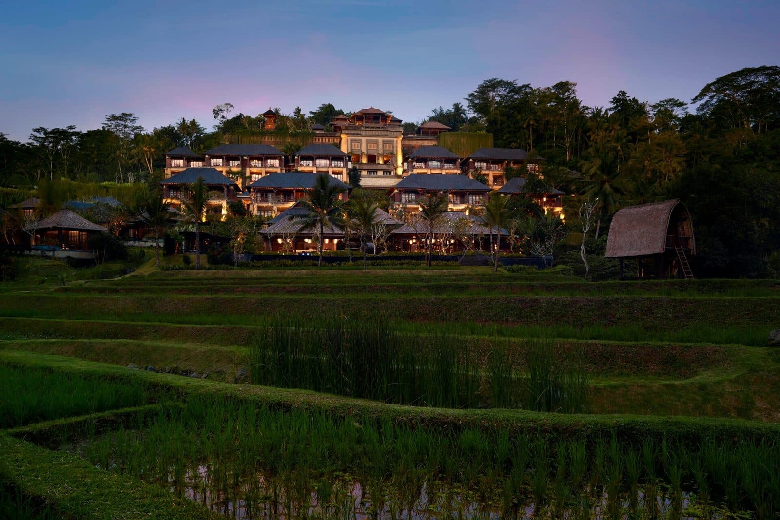 Mandapa at dusk: tiers of dark-roofed buildings above rice terraces, with a thatched rice barn on stilts to the right