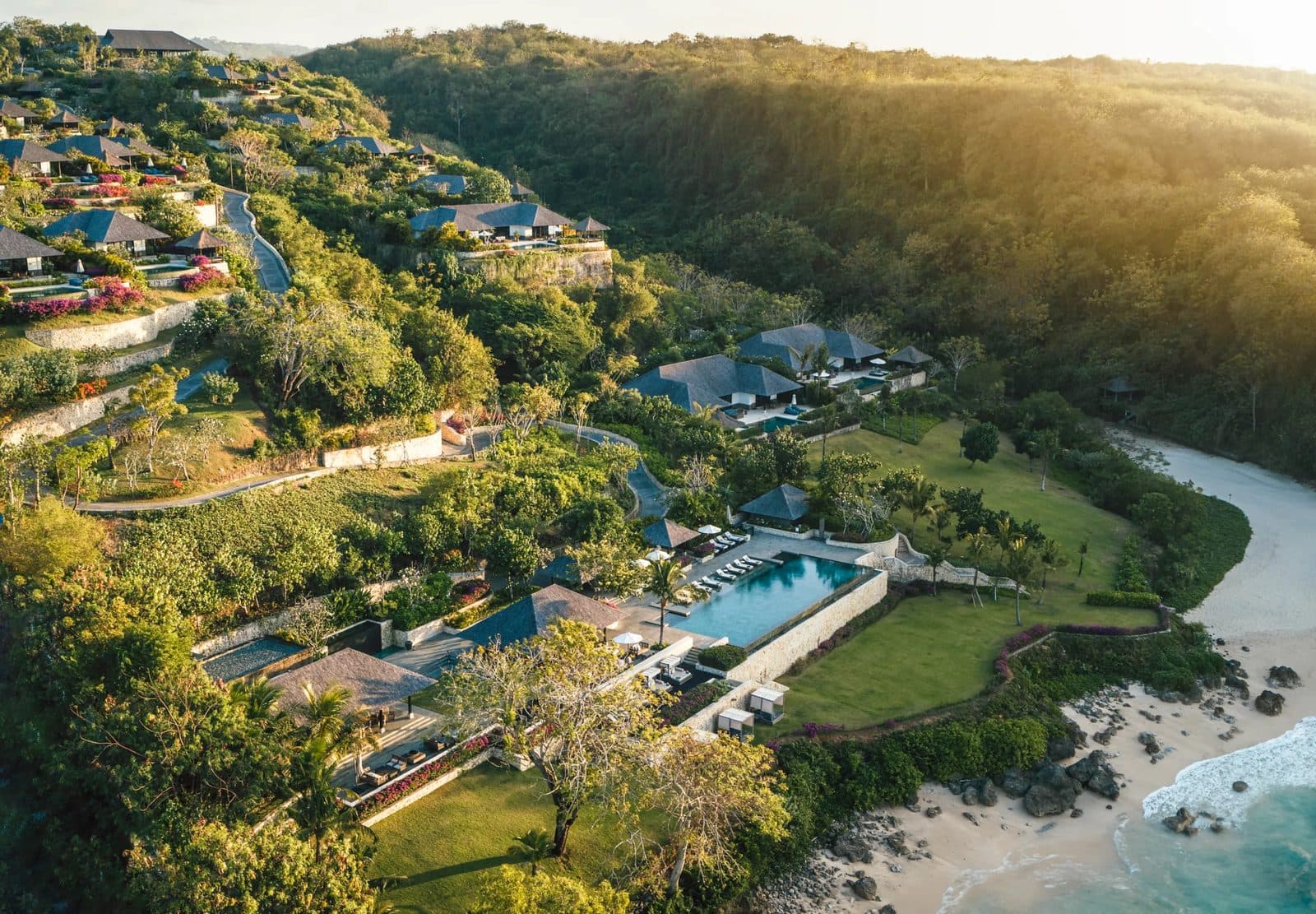 Aerial view of Raffles Bali: dark-roofed villas on a green slope, a pool beside the lawn and a beach in the bay below a wooded hill