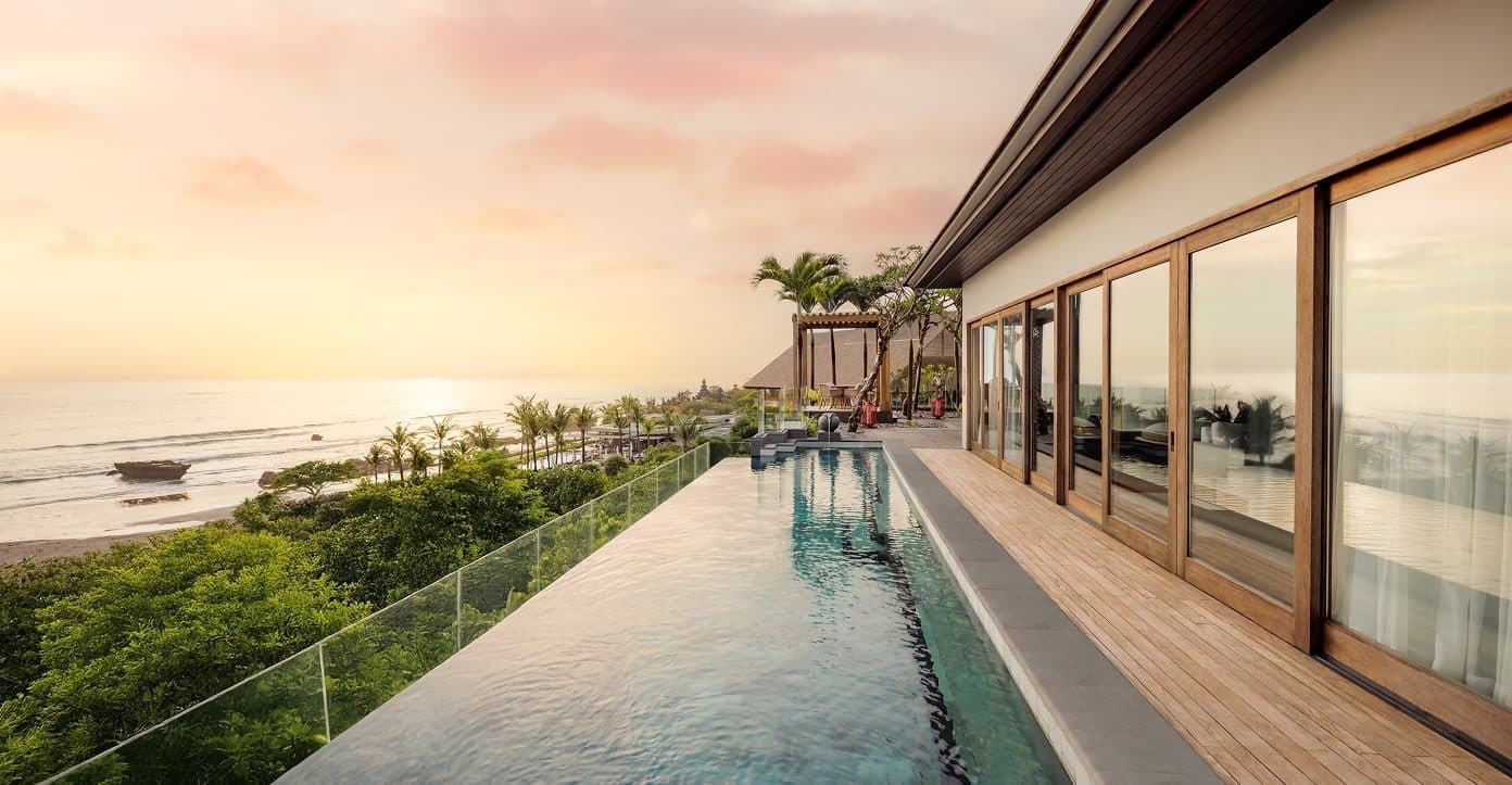Regent Bali Canggu penthouse infinity pool at sunset: a long pool beside a glass wall, with palms and a beach with a boat below