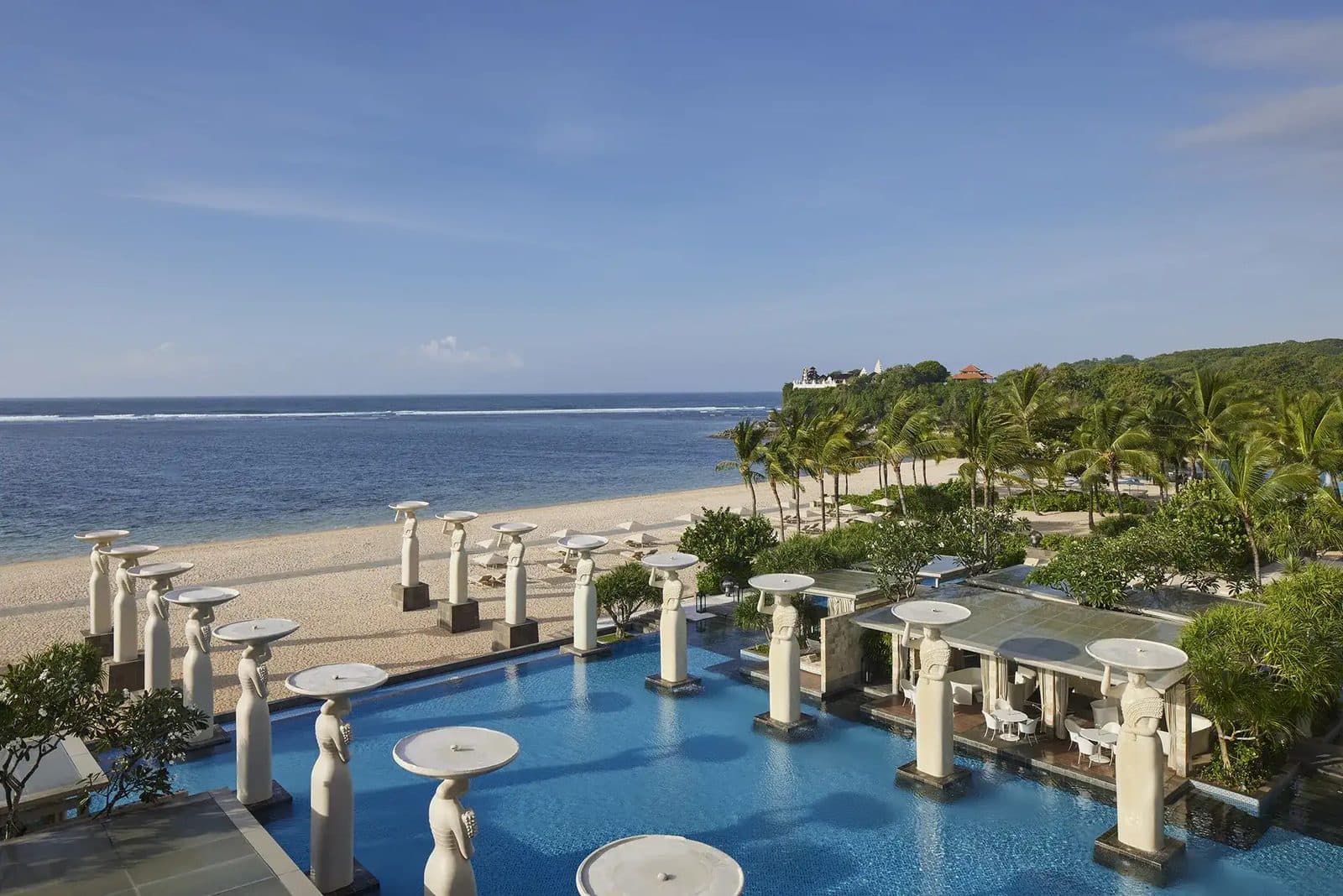 The Mulia's Oasis infinity pool: white statues holding bowls line the water, with Geger Beach, the ocean and a palm-covered headland beyond