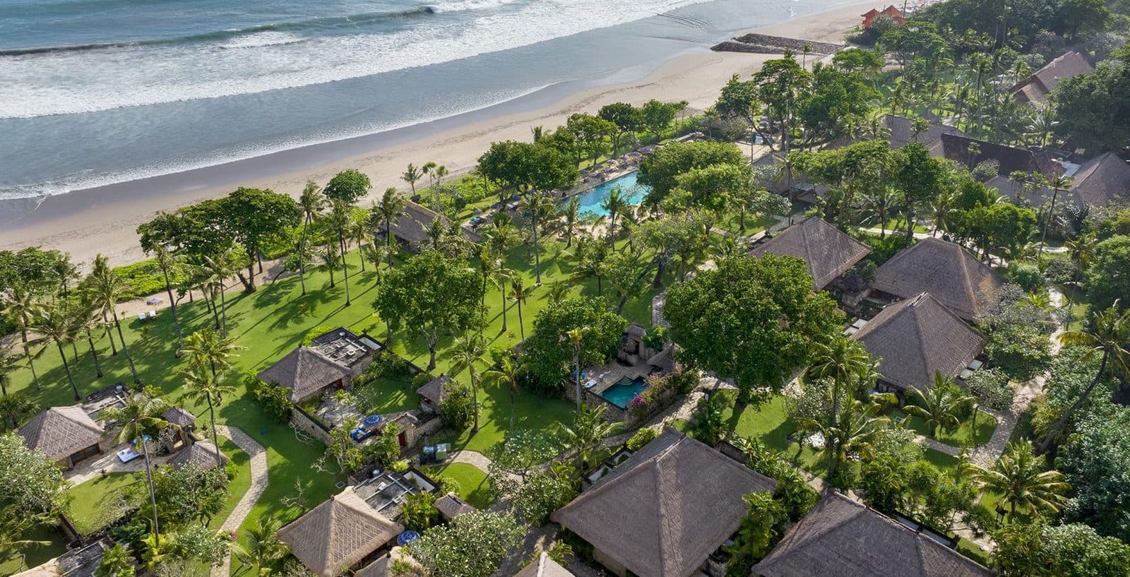 Aerial view of The Oberoi, Bali: thatched villa roofs among palms and lawns, a pool at the garden's edge and surf on Seminyak Beach