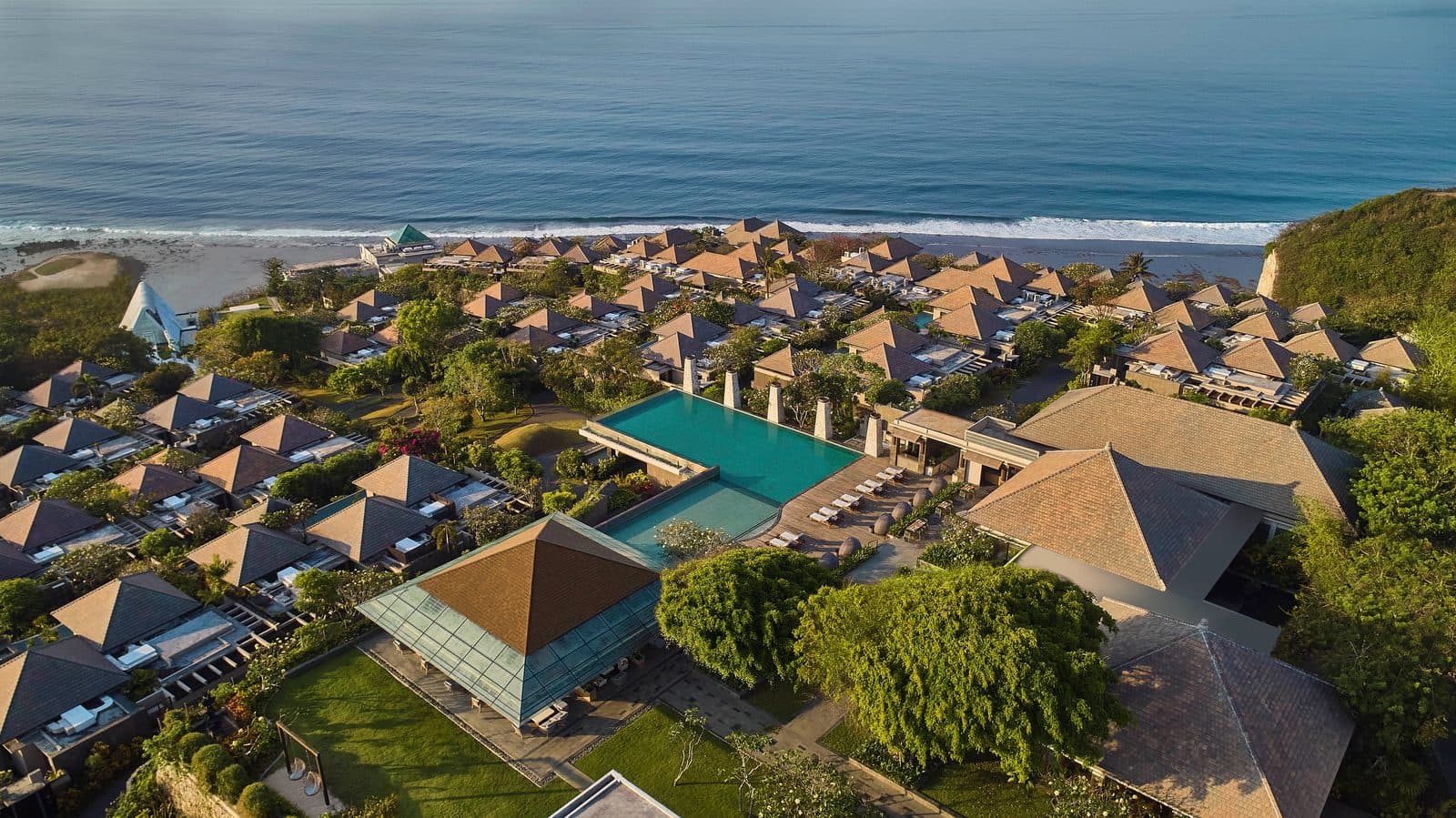 Umana Bali from above: terraces of villas with tent-shaped roofs descend towards Melasti Beach, with the main ocean-facing pool at the centre