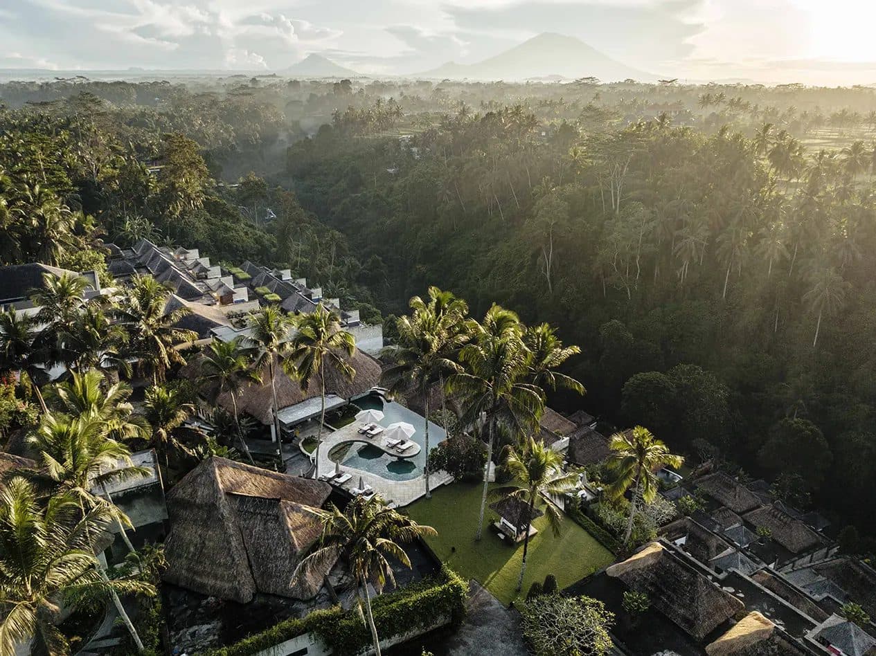 Aerial view of Viceroy Bali at sunrise: thatched villas, a circular pool with umbrellas and a lawn above the jungle valley, with a volcano in the distance
