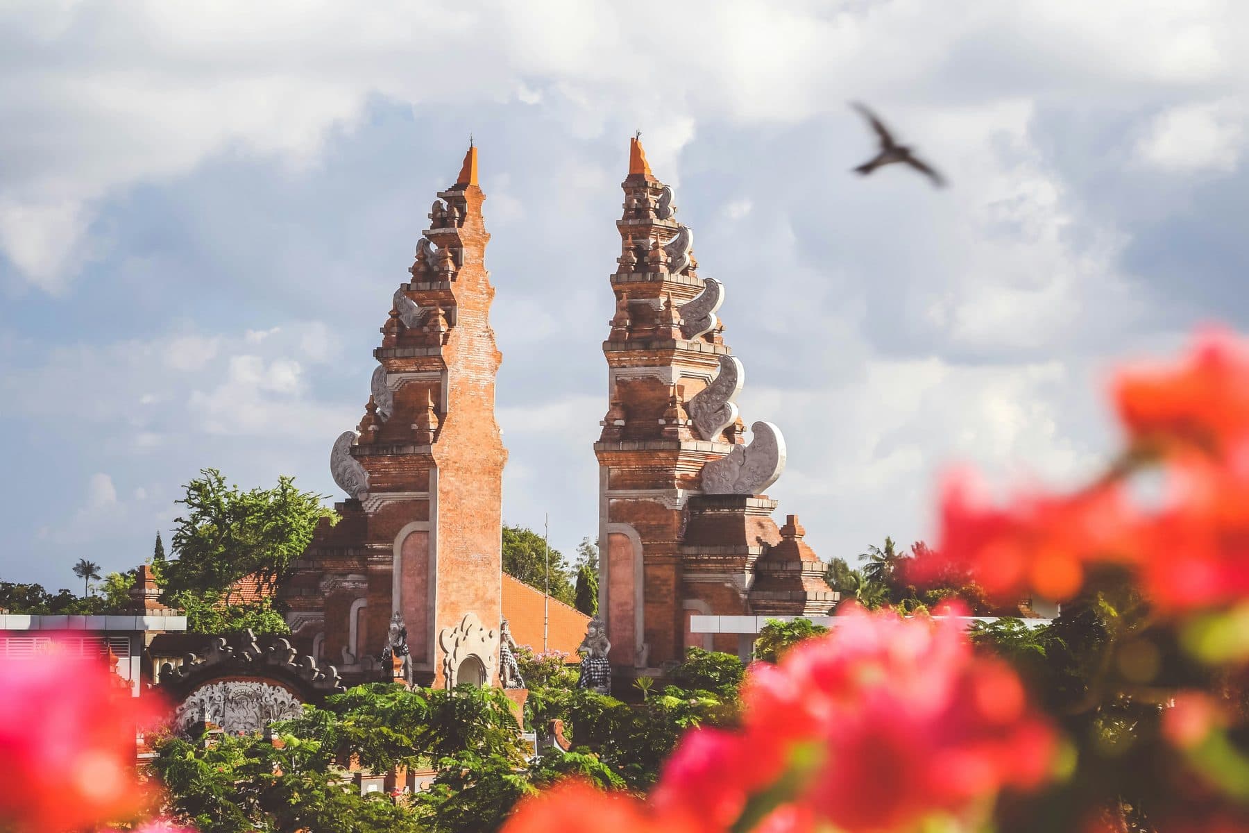 A red-brick split gate at a Balinese temple beneath cloudy skies, with blurred bougainvillea flowers in the foreground and a bird in flight