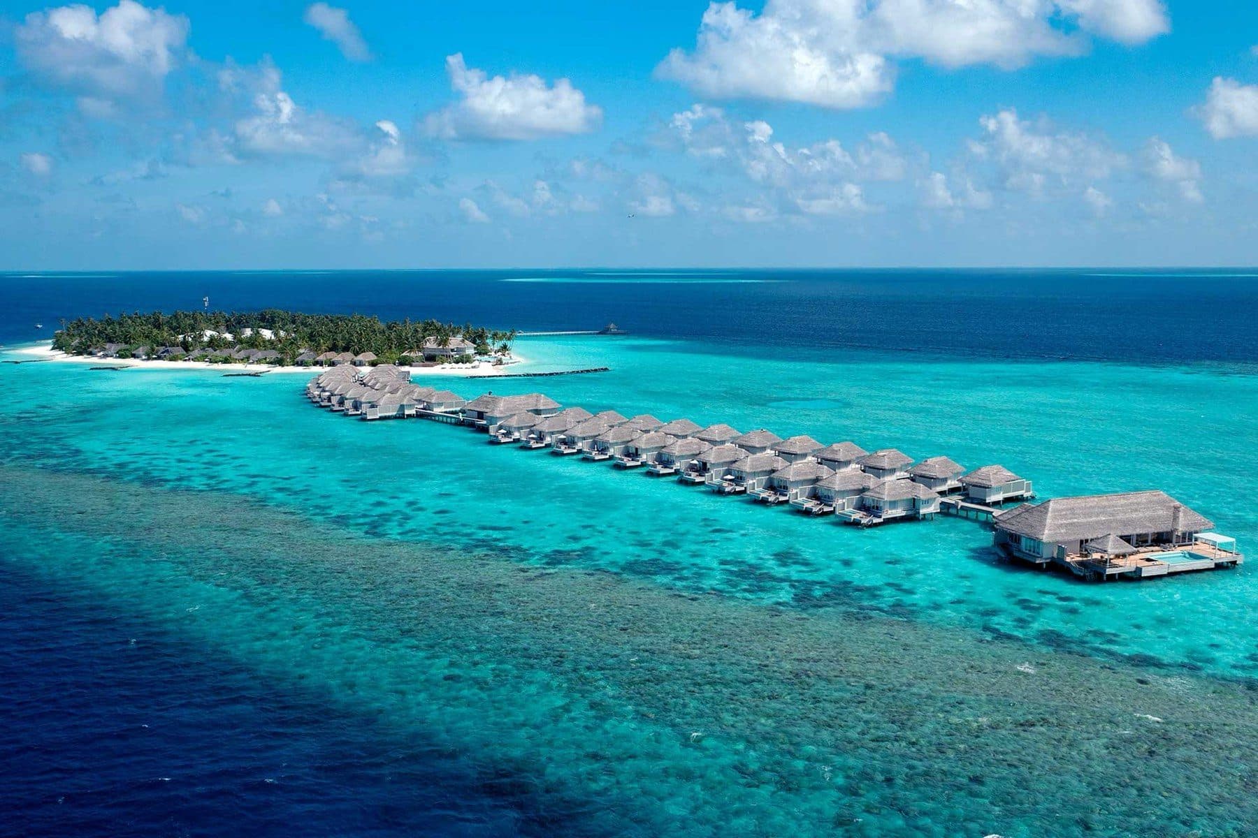 A long row of overwater villas on walkways above the reef ends at a large pool villa, with a palm-covered island in the distance; aerial view