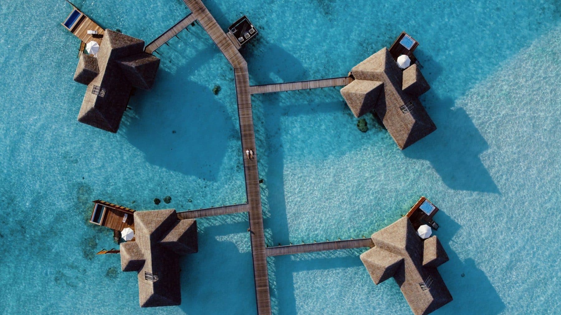 Direct overhead view of four thatched overwater villas with pools, connected by wooden walkways above the lagoon
