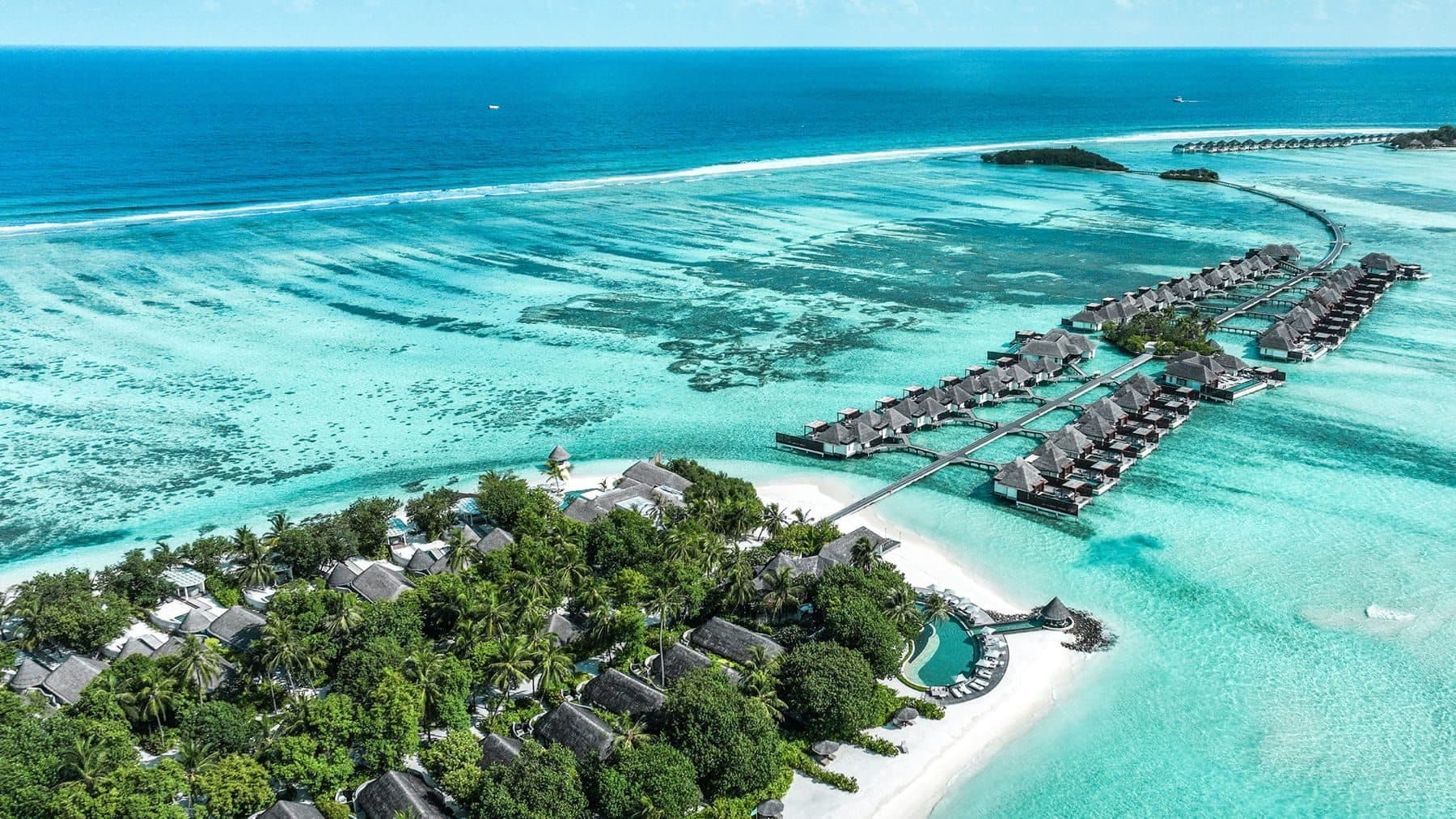 An island with villas among palms and rows of overwater villas on walkways above the lagoon, viewed from the air