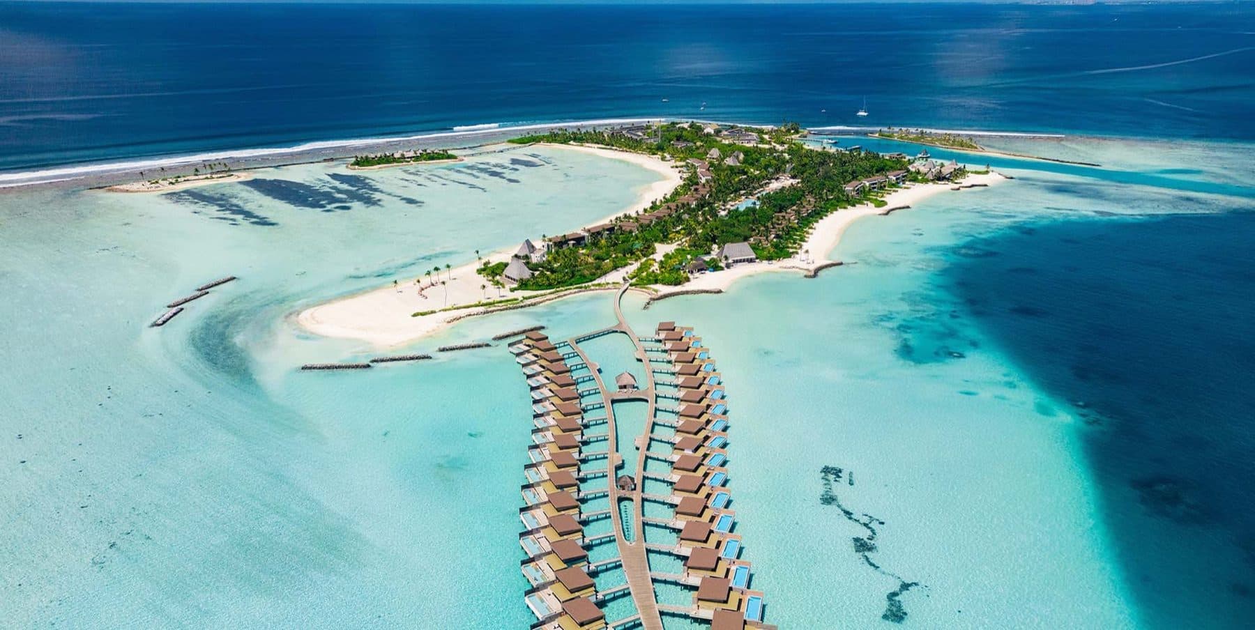 Aerial view of an island with beaches and palms and two rows of overwater pool villas along walkways