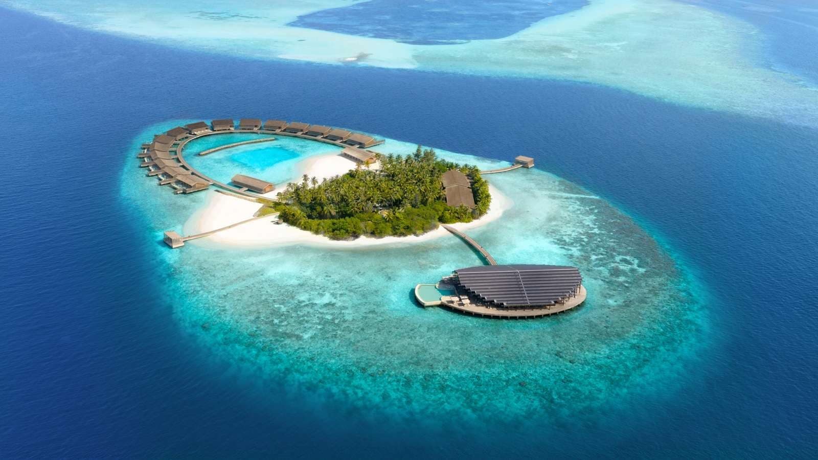 Aerial view of Kudadoo Island: an arc of overwater villas around a lagoon, a palm-covered island and a round pavilion with solar panels on the reef