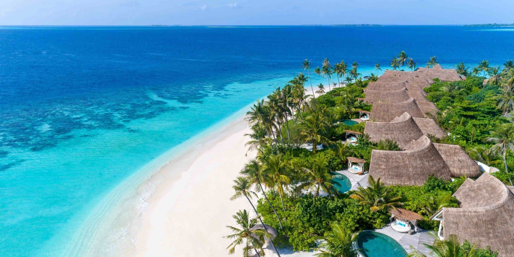 Thatched beach villas with pools along a white beach, viewed from the air
