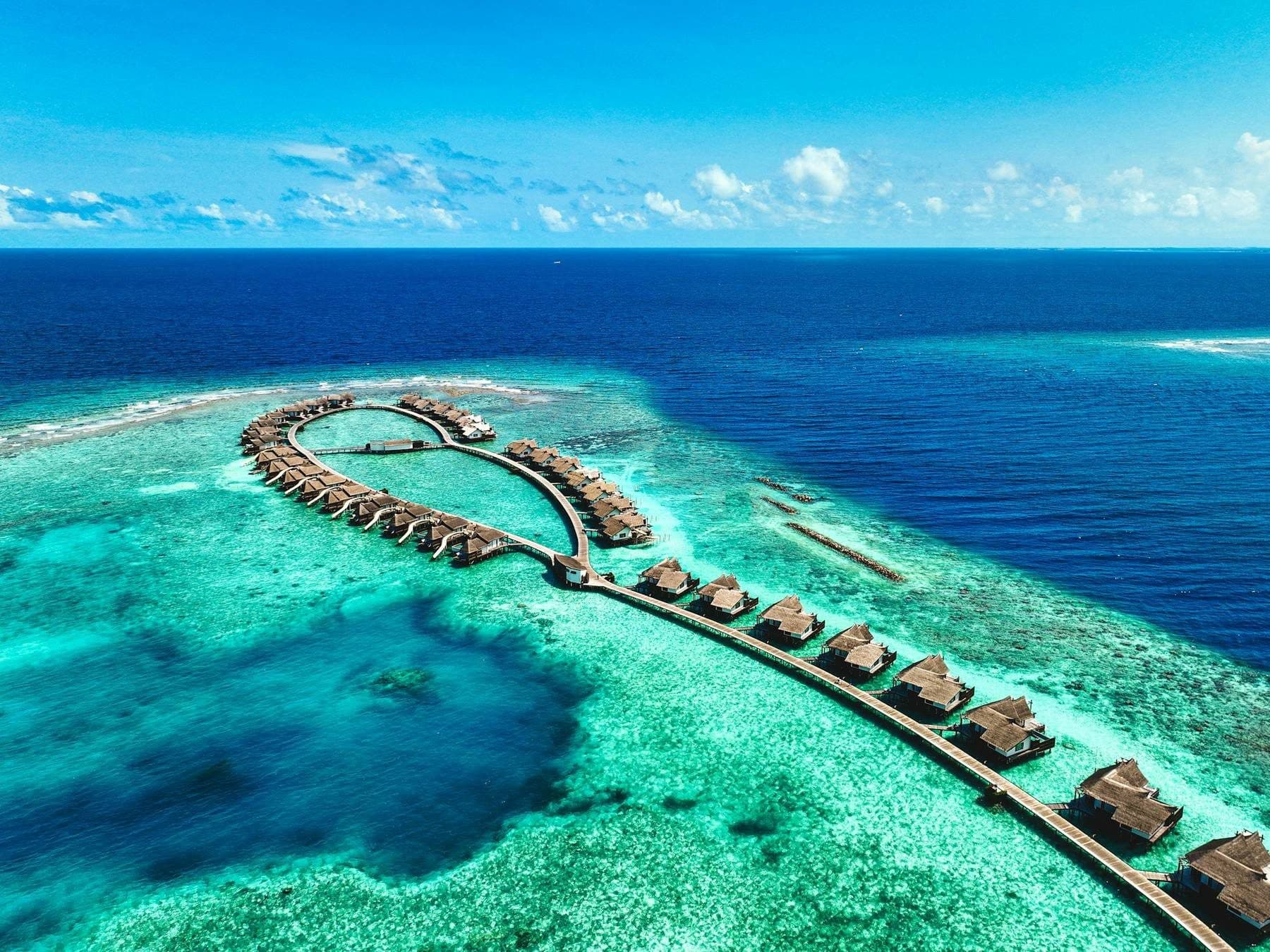 A loop and line of overwater villas on walkways above the reef, with the open ocean beyond, viewed from the air