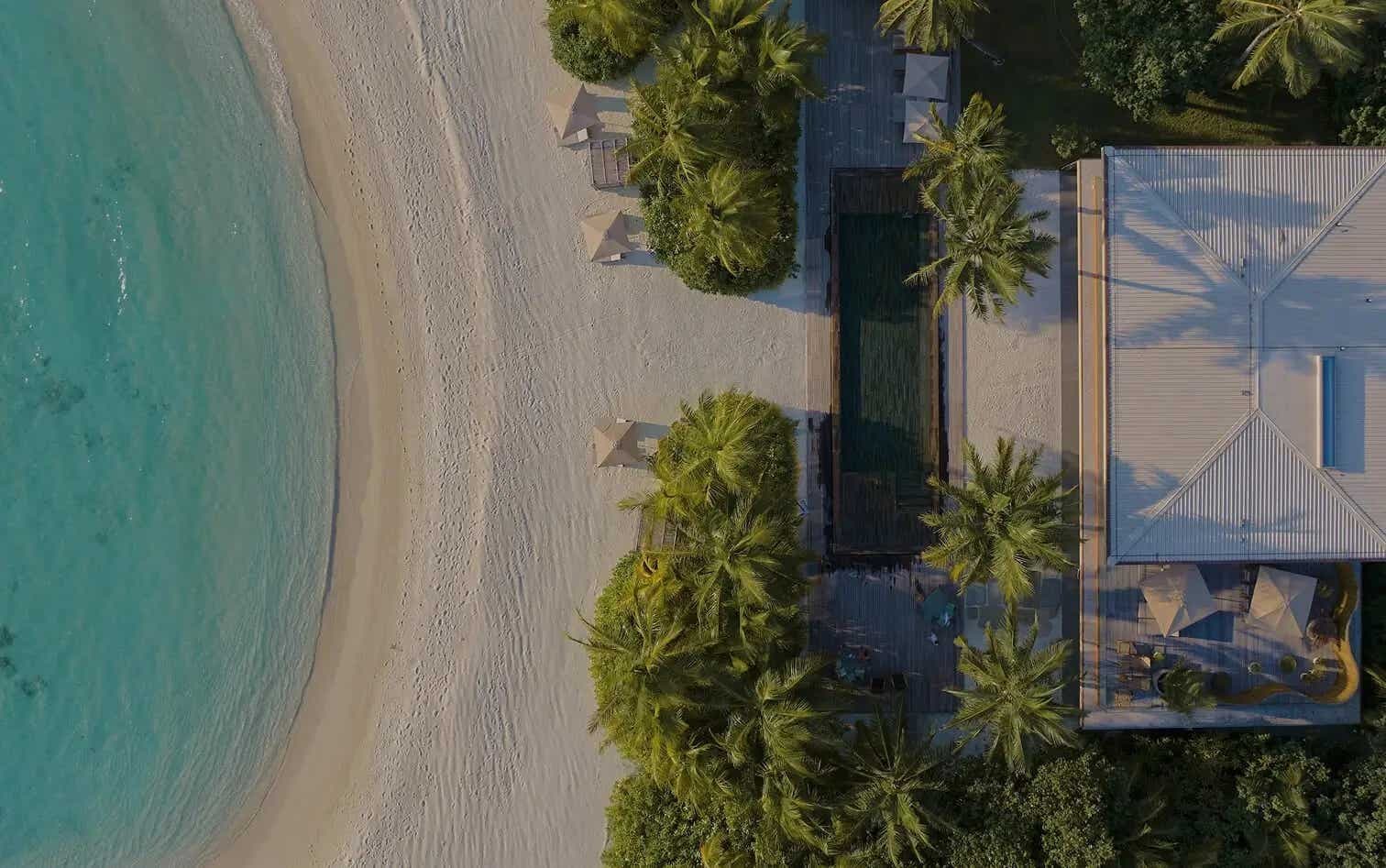 Aerial view of a villa with a long pool among palms, a white beach and turquoise water near shore