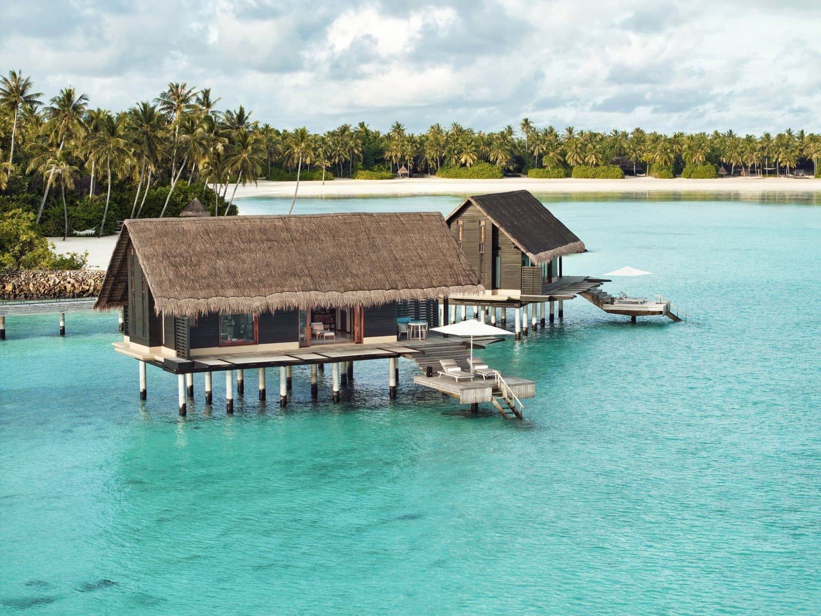 A thatched stilt villa above a turquoise lagoon; a white beach and palms behind it