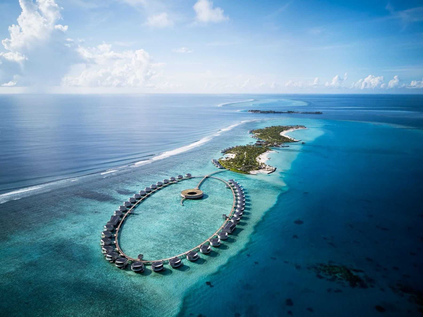 Aerial view of a ring of overwater villas with a round spa at its centre, and the resort's elongated islands beside the reef