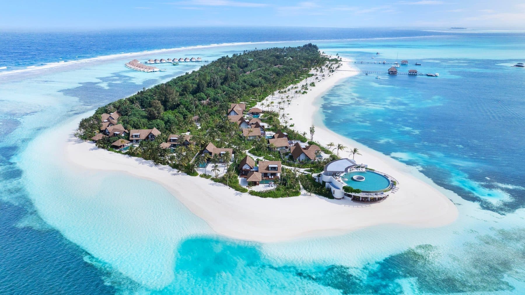 The tip of an island with villas among greenery, a round pool by the beach and a sand spit, viewed from the air