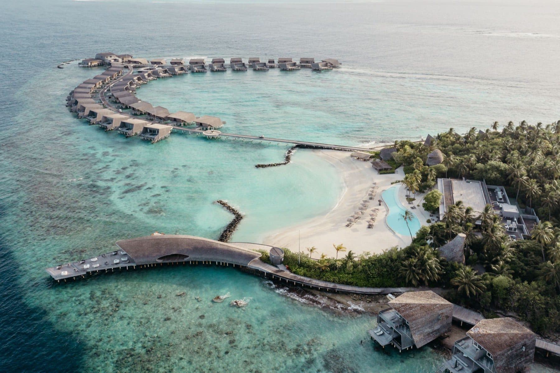 Aerial view of an arc of overwater villas, a beach with a pool and a curved bar building on stilts above the reef