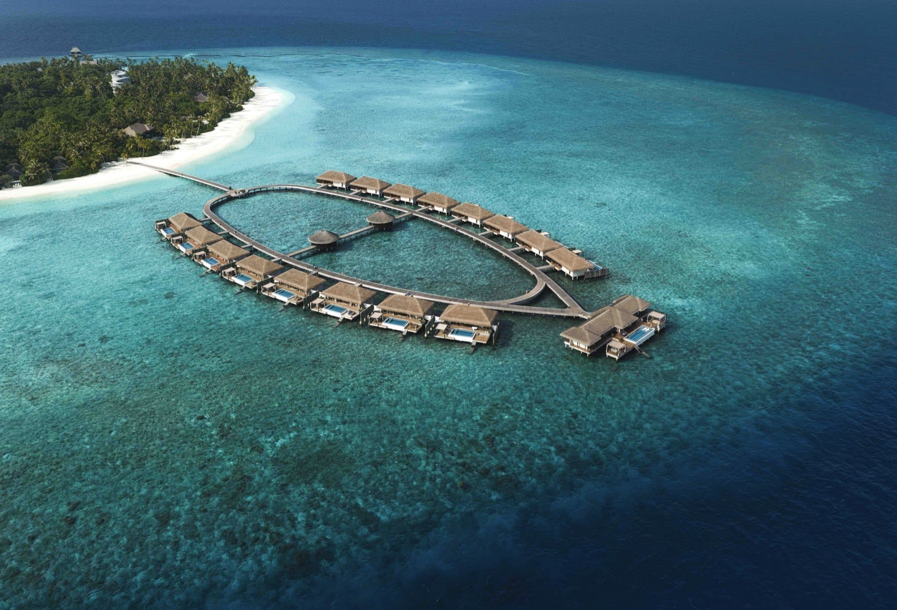Aerial view of a ring of overwater pool villas above the reef and the edge of the island with a beach and palms