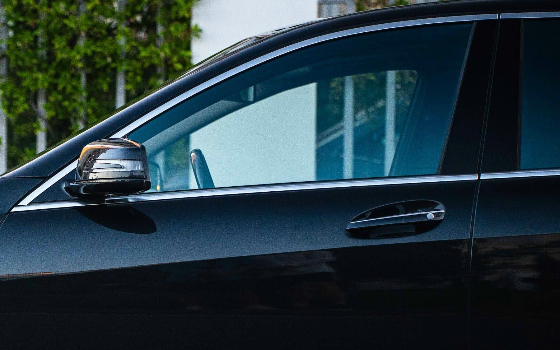 Close-up of a black saloon car door: side mirror, handle and tinted glass against a pale wall with greenery.
