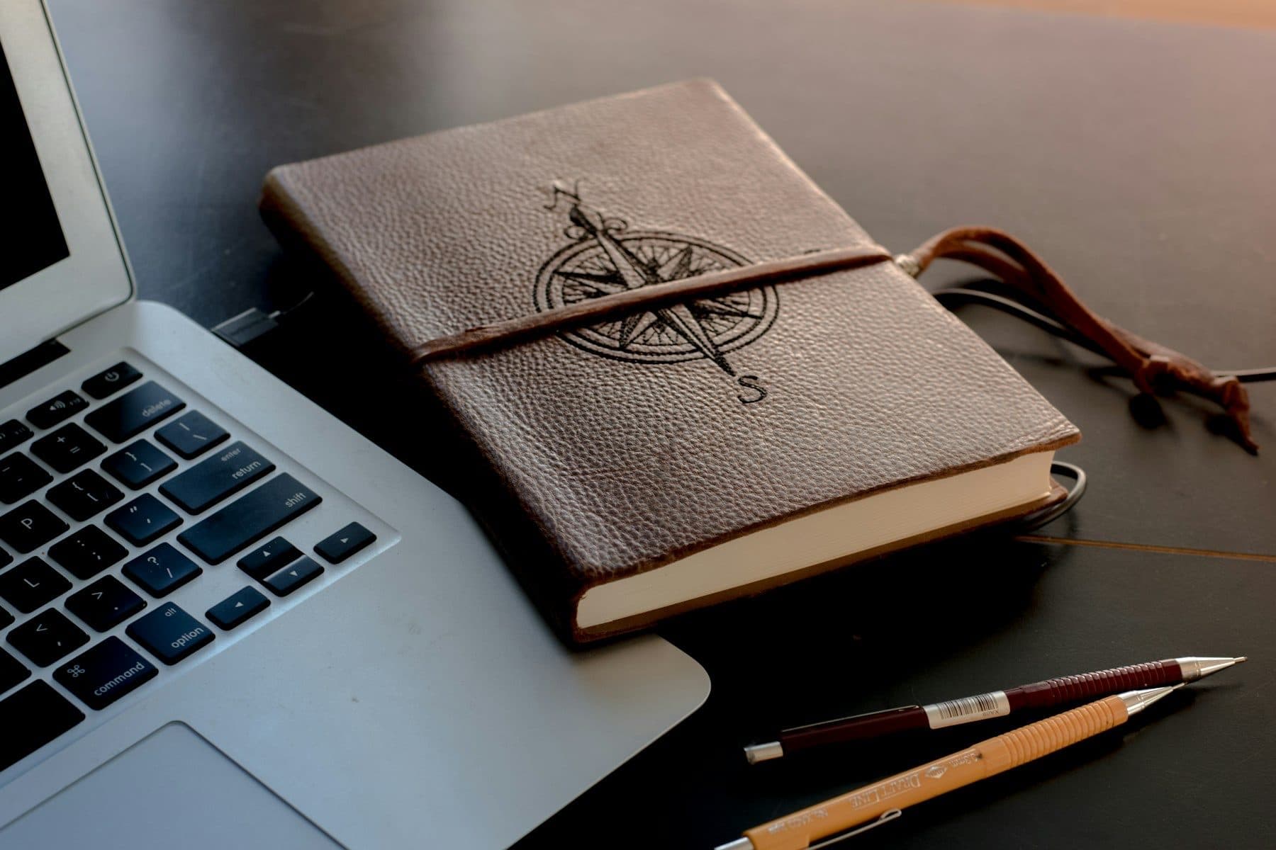 A leather notebook with a compass on the cover, a laptop and two mechanical pencils on a dark table.