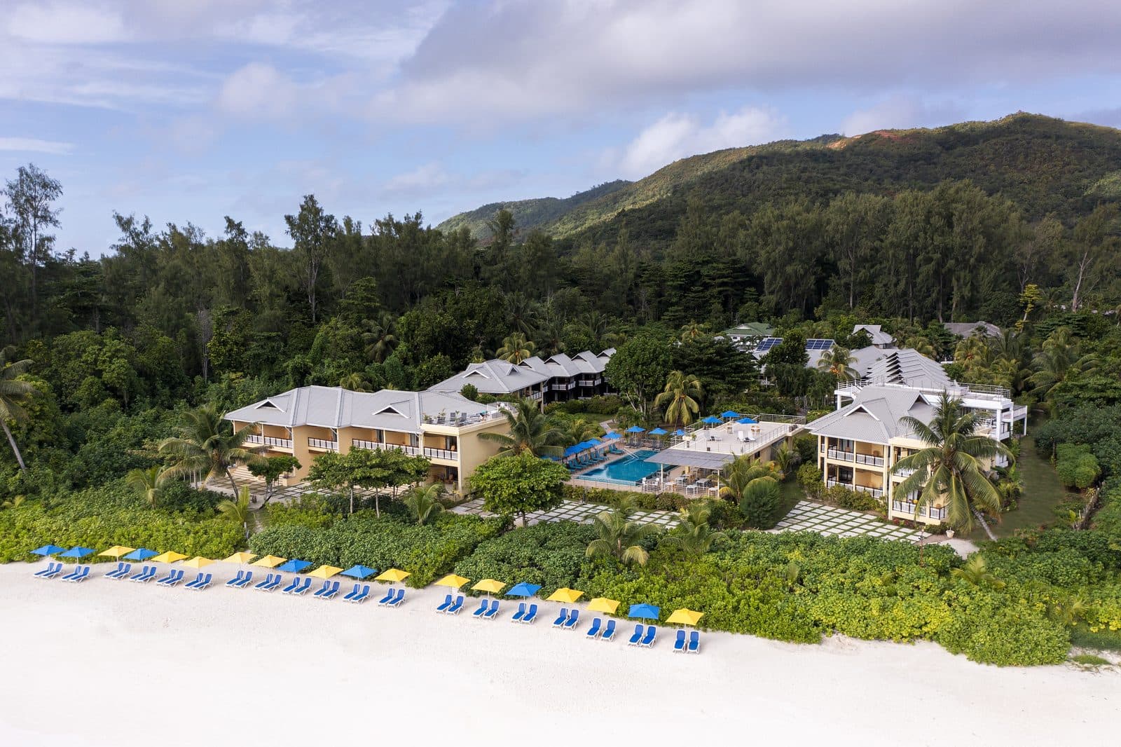Drone view of Acajou: grey-roofed buildings and a pool among greenery, blue and yellow umbrellas along a white beach, with Praslin's mountains behind