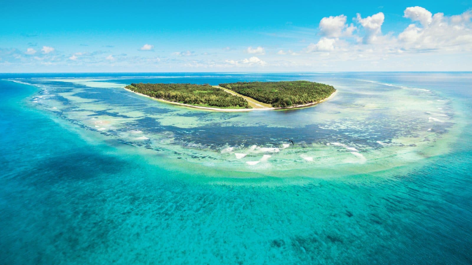 Aerial view of Alphonse Island: a green island with an airstrip through its centre, wide shallows and a turquoise lagoon around it