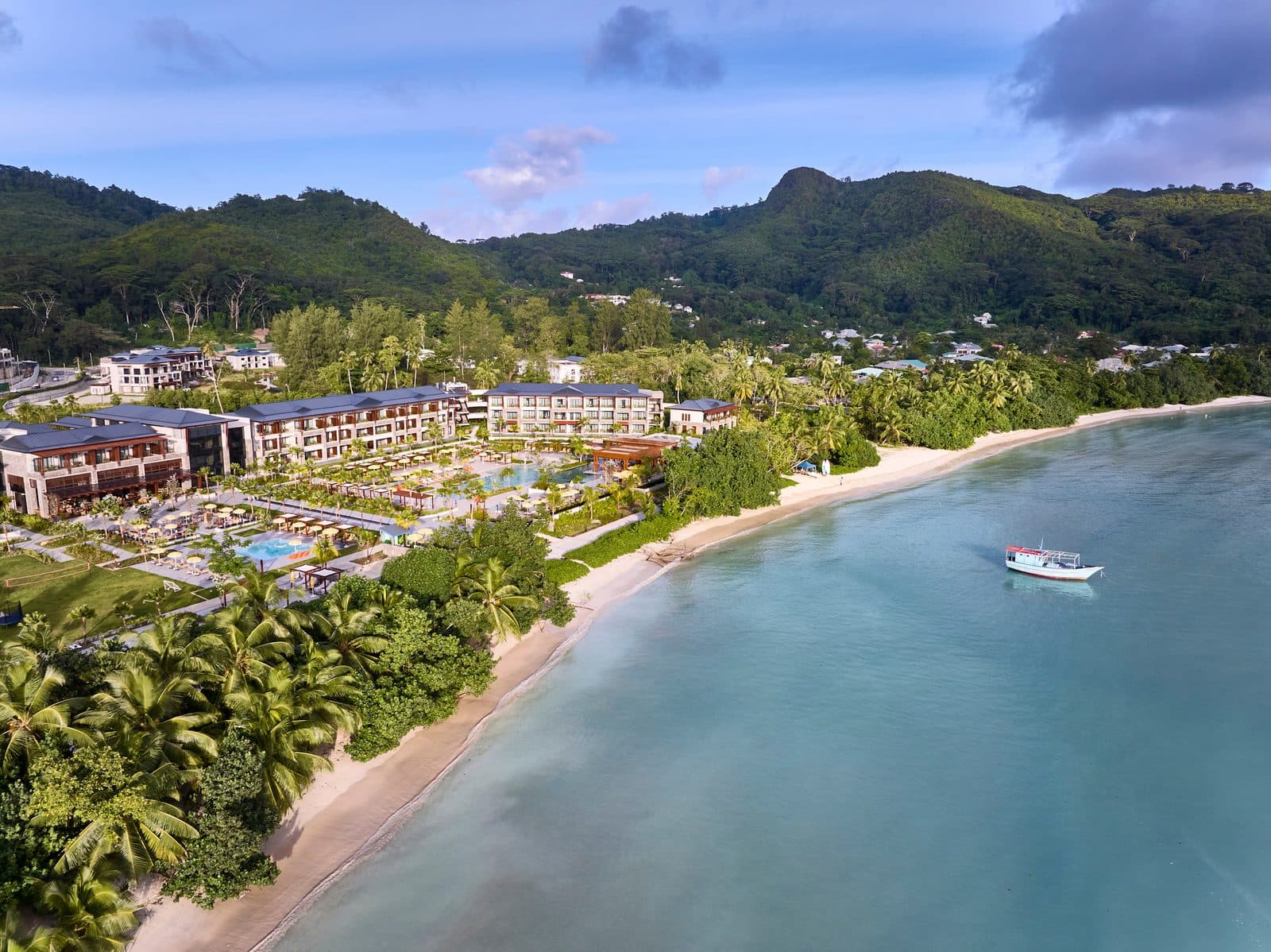 Aerial view of Canopy by Hilton Seychelles: buildings and pools among palms along Anse à la Mouche Bay's beach, a boat on turquoise water and Mahé's green mountains