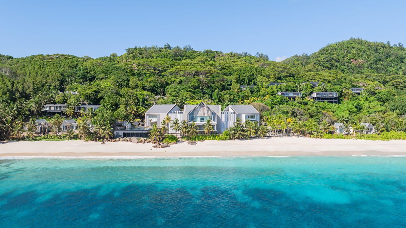 Anse Intendance Beach seen from the sea: white villas with pitched roofs among palms at the sand's edge, hilltop villas further up the jungle slope and turquoise water in the foreground