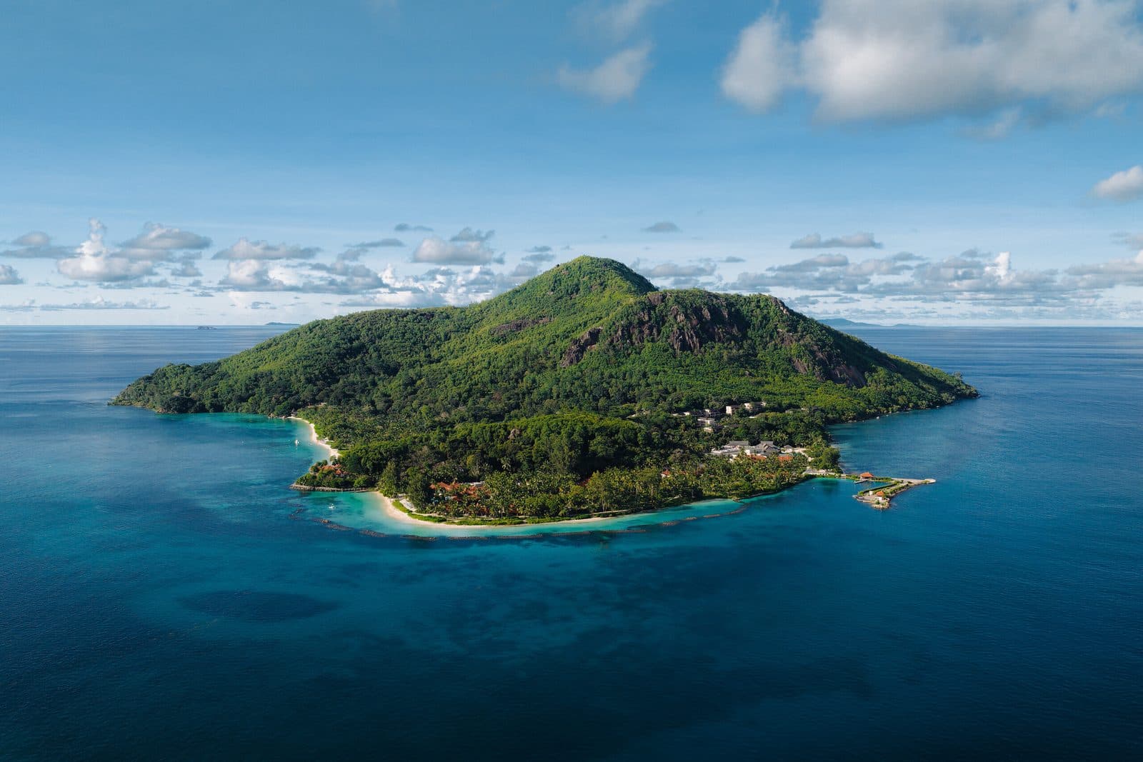 Aerial view of Sainte Anne Island: a green mountain, white beach with resort buildings among trees and a jetty in the blue ocean