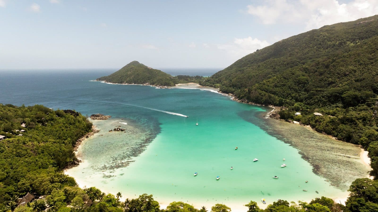 Aerial view of the bay beside the resort: a turquoise lagoon with boats, a reef along the shores, wooded slopes and a headland at the opening to the ocean
