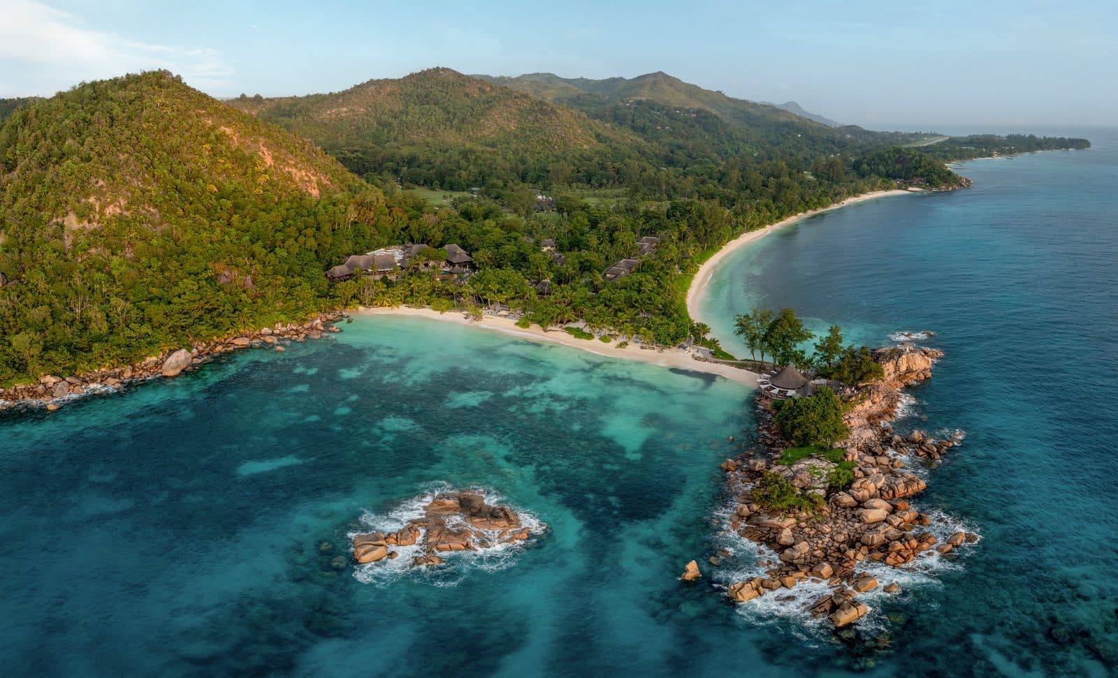 Aerial view of the Anse Kerlan bays: resort roofs amid forest at the foot of mountains, a white beach, a granite headland between two beaches and turquoise water