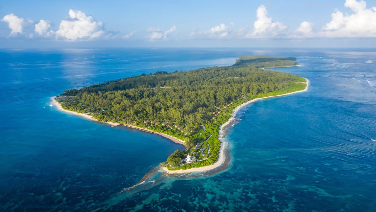 Aerial view of Desroches Island: an elongated wooded island with white beaches, a lighthouse on the headland and a reef in turquoise water