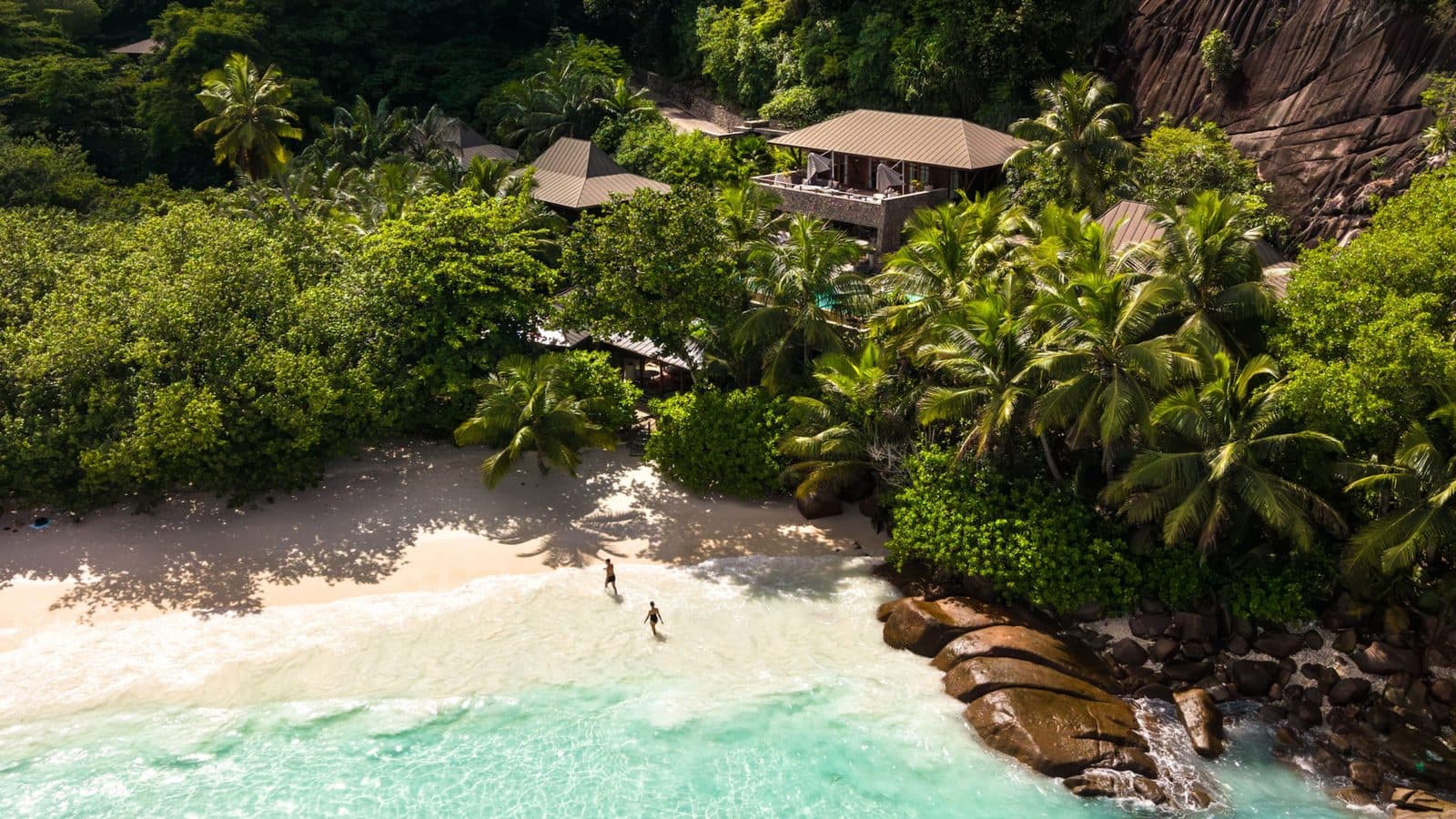 Aerial view of Petite Anse Beach: white sand, turquoise water, granite boulders and villa roofs among hillside palms