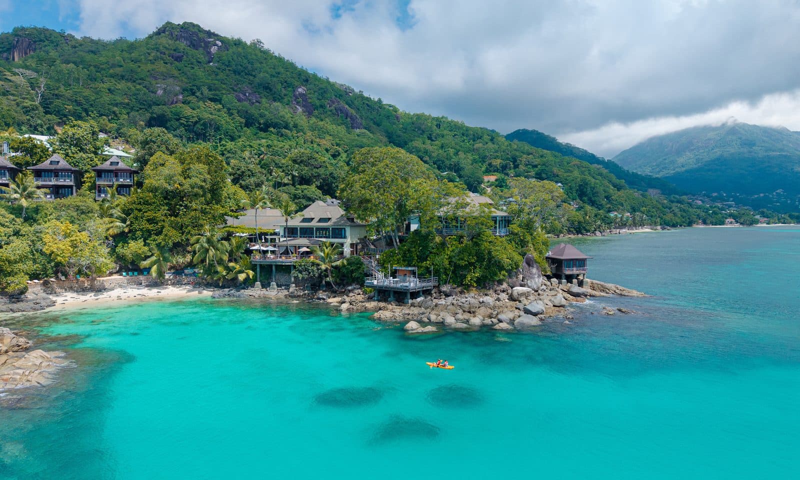 Aerial view of Hilton Northolme: villas on a green slope above granite rocks, a turquoise bay and a kayak near shore