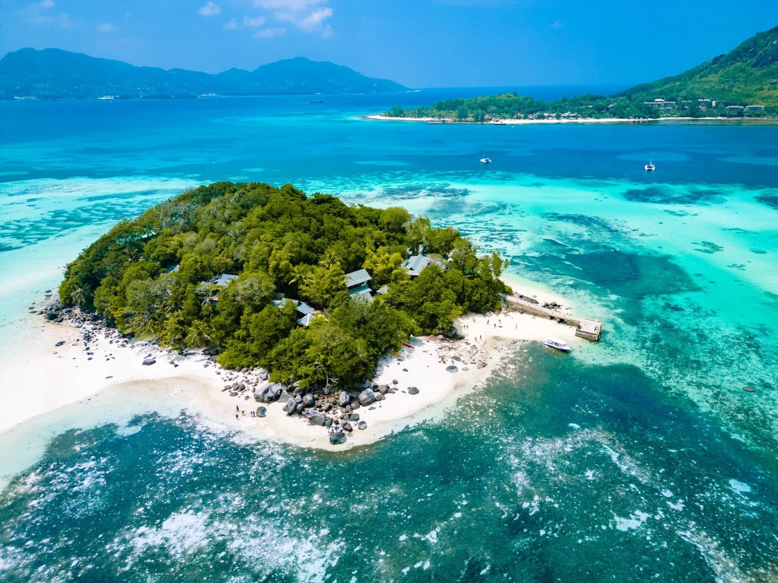 Aerial view of Round Island: a green hill with villa roofs, granite boulders, white sand, a jetty with a boat and turquoise shallows, with mountains in the distance