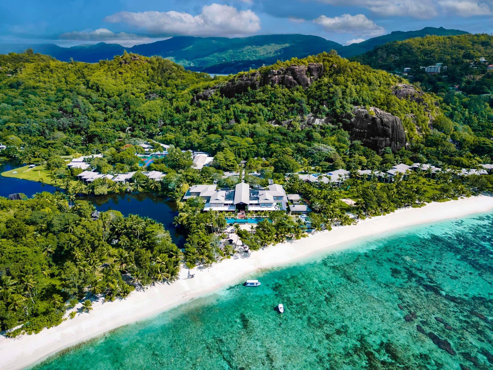 Aerial view of Kempinski Seychelles: buildings among palms between the lagoon and Baie Lazare beach, a reef in turquoise water and wooded mountains with granite rocks
