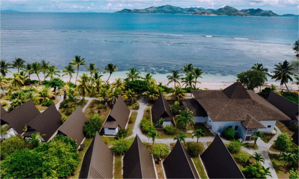 Aerial view of La Digue Island Lodge: triangular chalet roofs among palms, the white sand of Anse Réunion Beach and Praslin across the strait