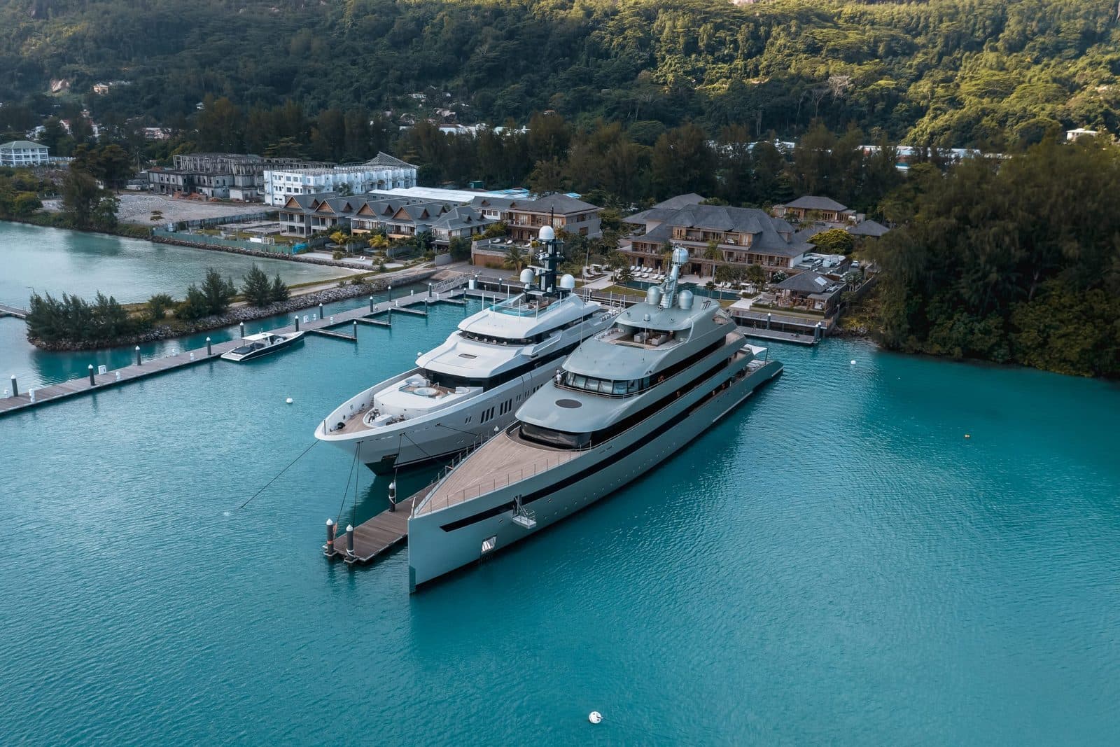 Aerial view of L'Escale marina: two superyachts beside the pontoon, with hotel roofs and Mahé's green hills behind