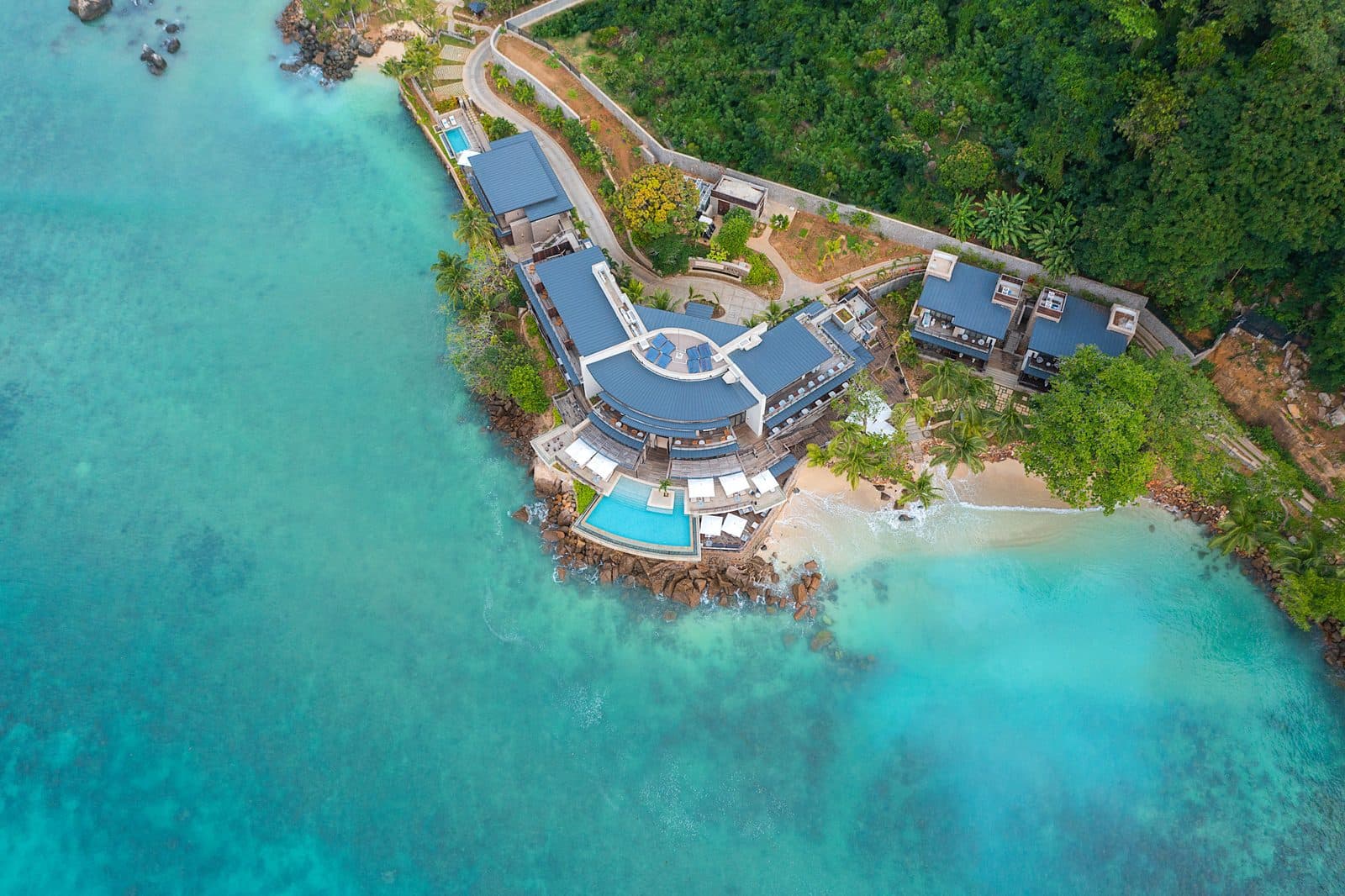 Aerial view of Mango House: blue-roofed buildings on a rocky headland, a pool at the water's edge, a small beach and a turquoise bay