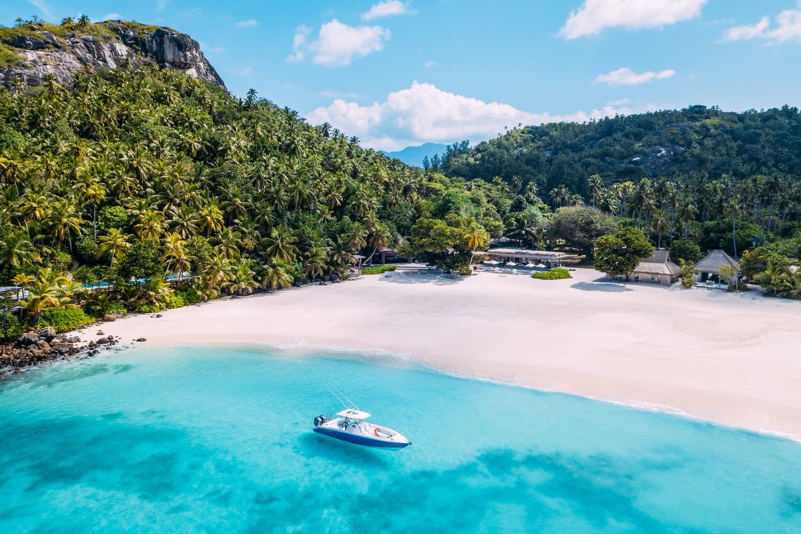 Aerial view of North Island's East Beach: a speedboat on turquoise water, white sand and villa roofs among palms beneath a granite cliff