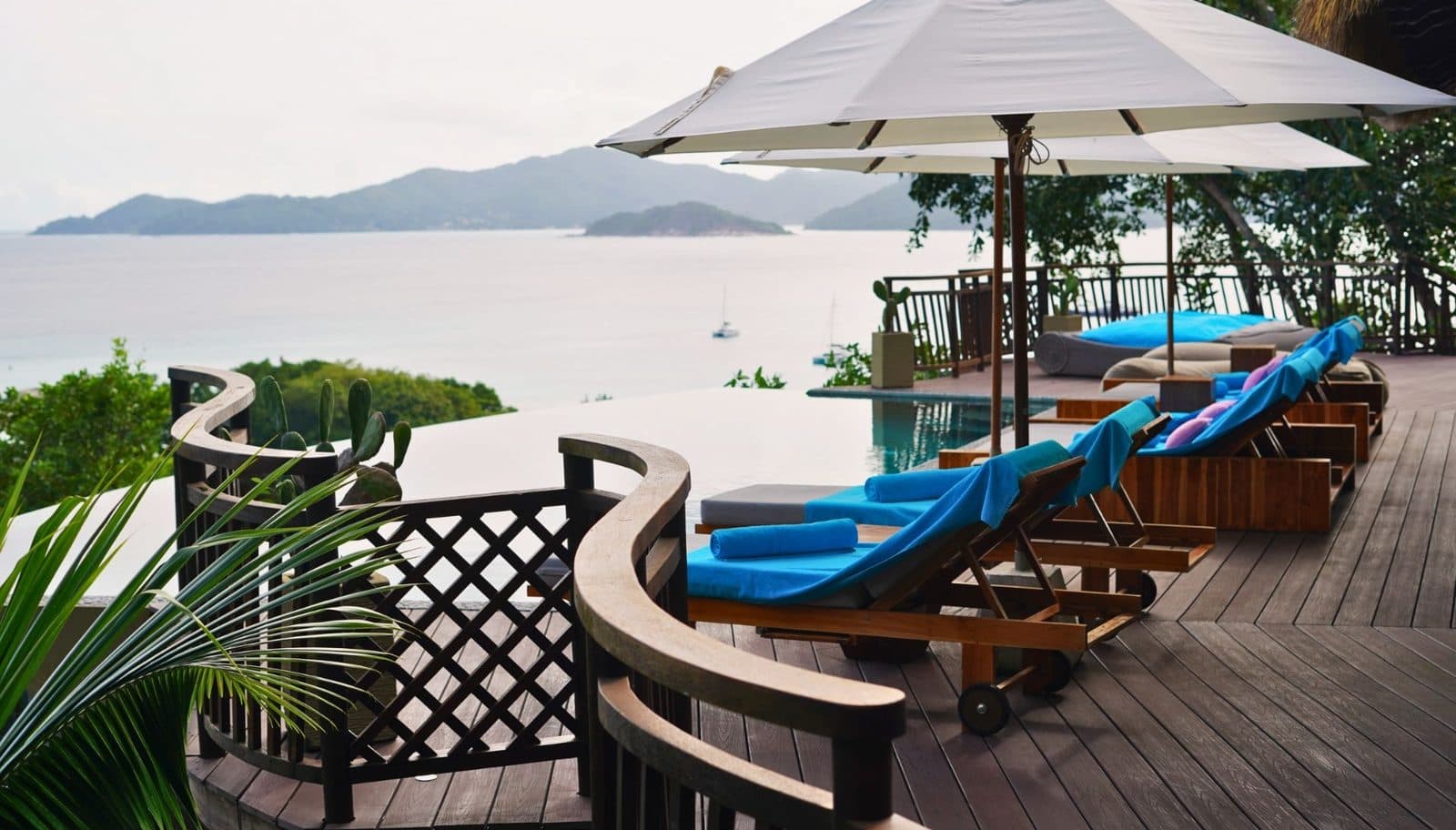 An infinity pool on a wooden terrace with sun loungers beneath umbrellas. Beyond are the sea, sailing boats and the mountains of a neighbouring island