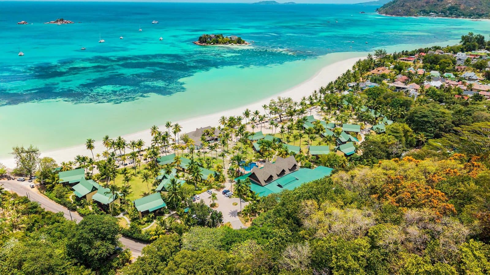 Drone view of Paradise Sun: green-roofed bungalows among palms along white Côte d’Or Beach, turquoise water and St Pierre islet