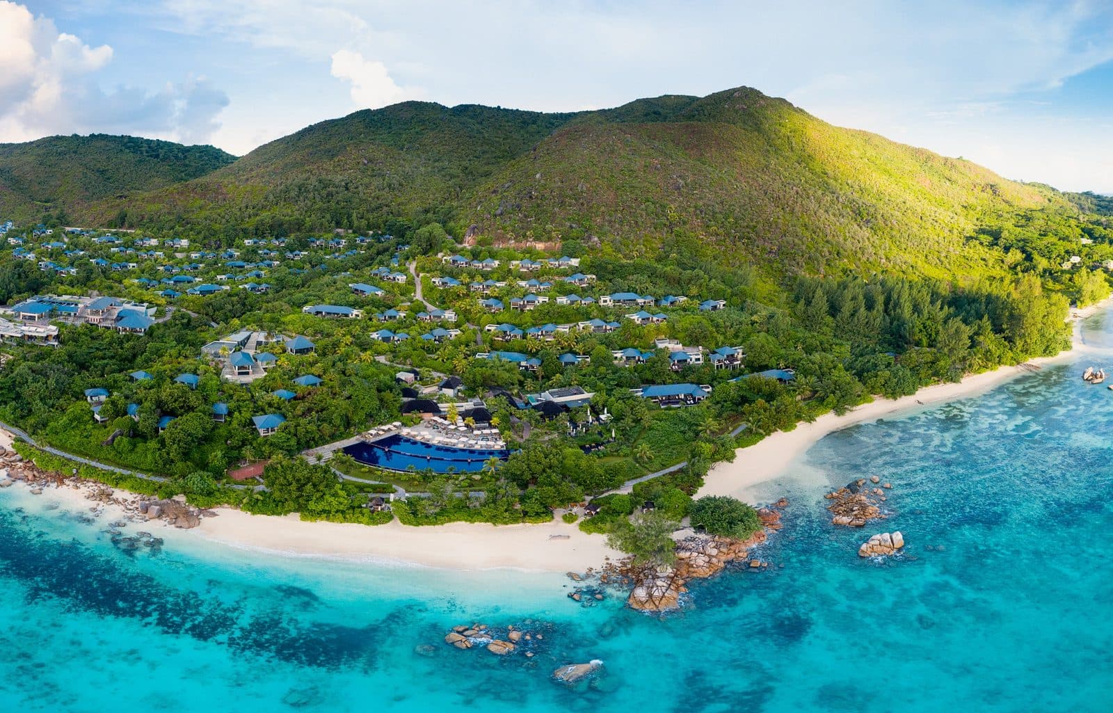 Aerial view of Raffles Seychelles: blue-roofed villas on a green mountainside, a large seaside pool, white beach and turquoise water with granite rocks