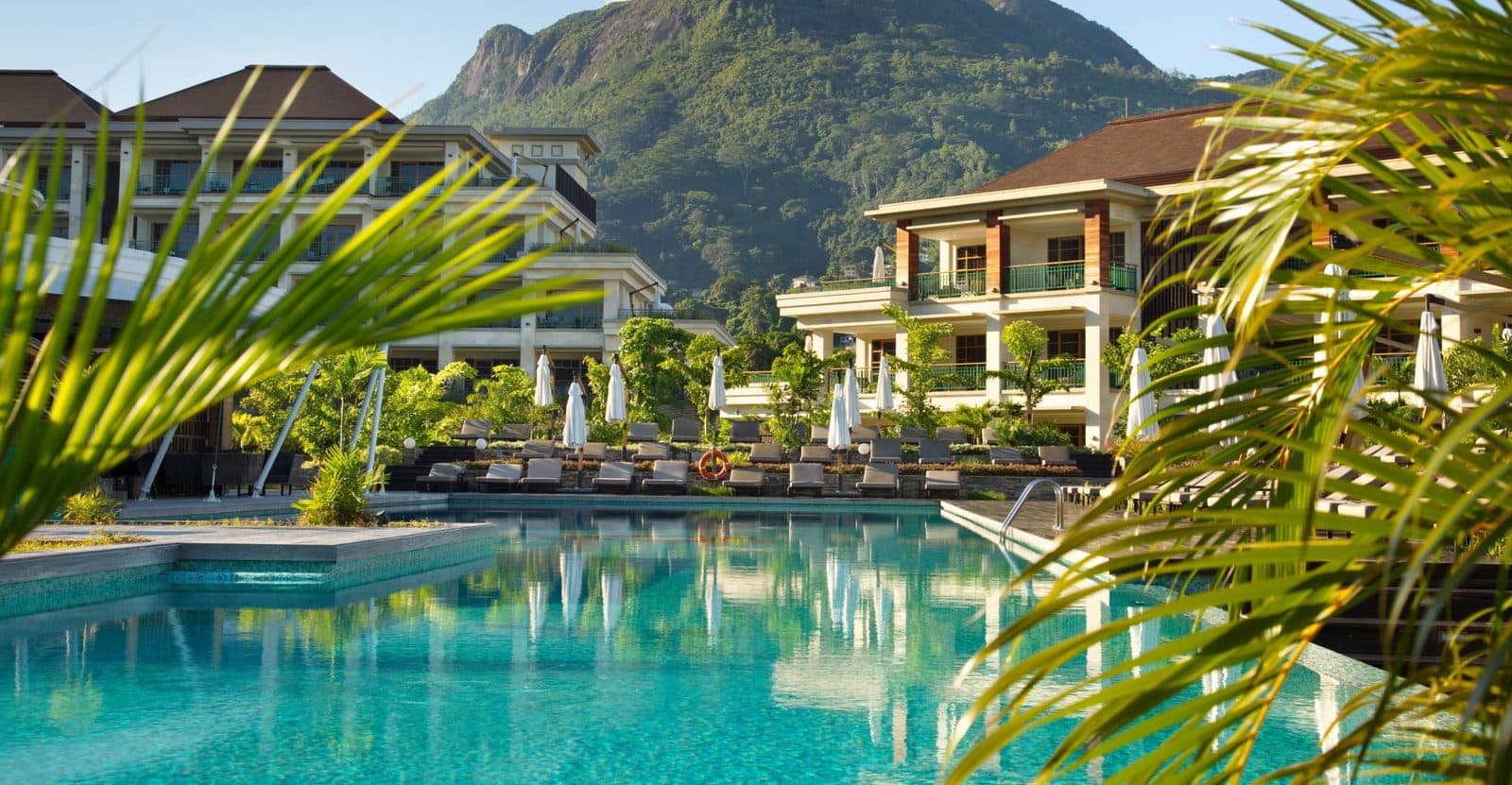 Savoy's large pool framed by palm leaves: sun loungers beneath umbrellas, hotel buildings with balconies and a green mountain above